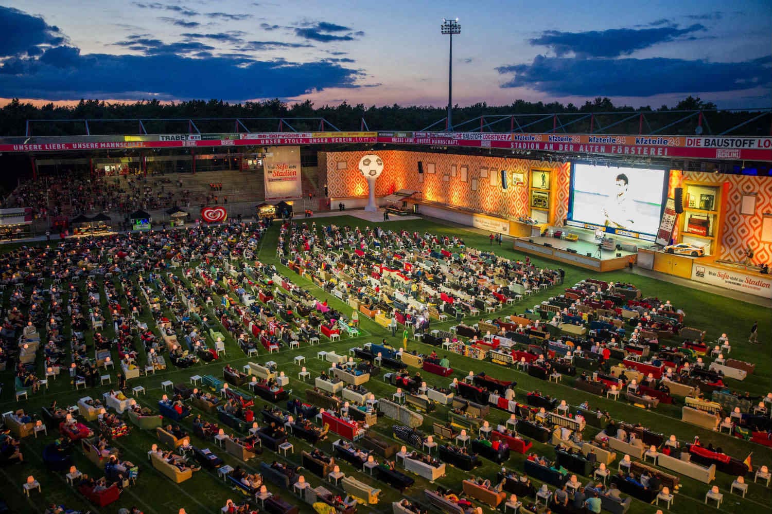 Huge TRUE dvLED screen at a stadium public viewing at dusk with seating on the pitch