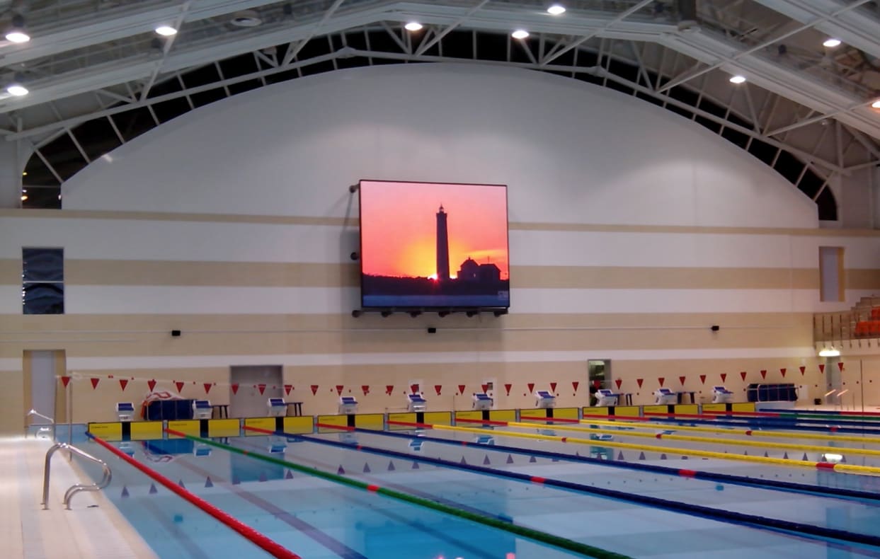 Indoor competition swimming pool with a TRUE dvLED screen on the wall showing a sunset scene