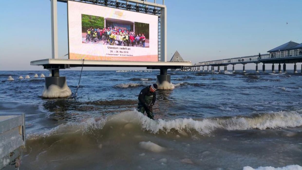Large TRUE dvLED screen on a seaside pier while a man wades through the breaking waves